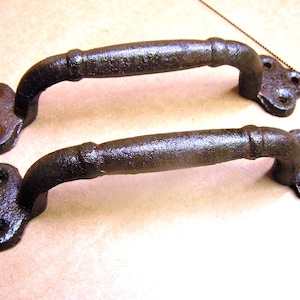 May include: Two rustic, dark brown cast iron handles. Each handle has a curved grip and decorative, four-hole mounting plates. The handles are likely for a cabinet or drawer, adding a vintage touch.