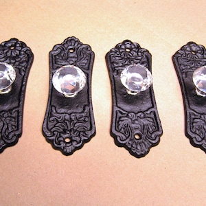May include: Four black metal wall hooks, each featuring an ornate, rectangular backplate with intricate scrollwork and a single screw hole at the top and bottom. A clear, faceted, round glass knob is centered on each black metal backplate.