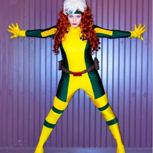 May include: A woman dressed as a superhero, wearing a yellow and green costume with a white wig and red hair. She is standing with her arms outstretched in front of a purple wall.