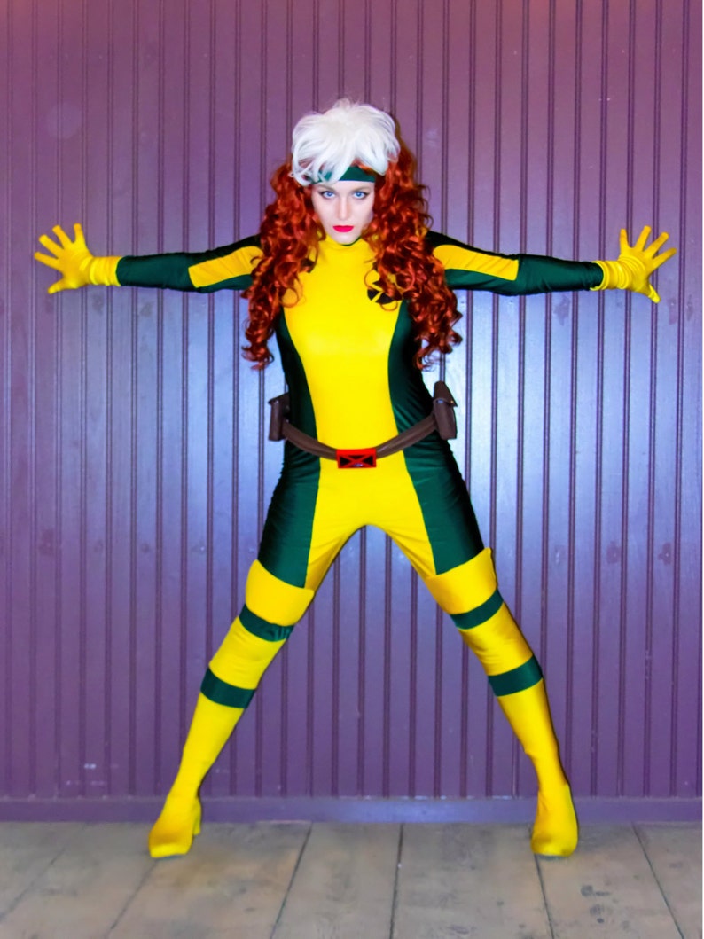 May include: A woman dressed as a superhero, wearing a yellow and green costume with a white wig and red hair. She is standing with her arms outstretched in front of a purple wall.