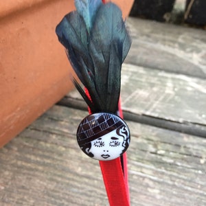 May include: Red headband with a black feather and a white button featuring a black and white illustration of a woman wearing a hat.
