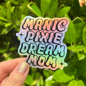 May include: A holographic sticker with the text "Manic Pixie Dream Mom" in a rainbow gradient. The sticker has a glittery finish and features two small stars.