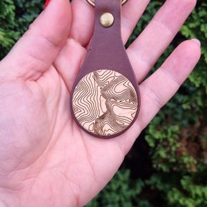 May include: A brown leather keyring with a round wooden charm featuring a topographic map design. The keyring has a brass key ring.