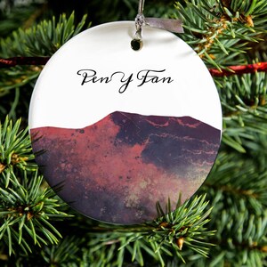 May include: A white ceramic Christmas bauble featuring a red and purple mountain scene with the text "Pen y Fan" in black cursive.