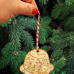 Pode incluir: Um enfeite de Natal de madeira em forma de mapa de uma cidade. O enfeite está pendurado em uma corda vermelha e branca em uma árvore de Natal verde. O mapa apresenta nomes de ruas como "Greenfield House", "Priory Sch" e "Cartmel".