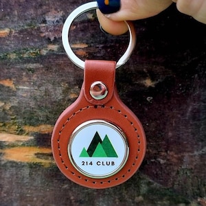 May include: A brown leather keyring with a silver ring and a round metal tag. The tag has a white background with a green mountain design and the text "214 CLUB".