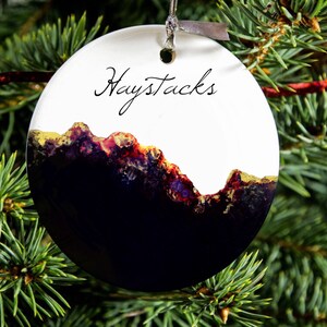 May include: A white round Christmas bauble with a black and gold abstract design and the text "Haystacks" written in black cursive.