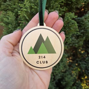 May include: A circular wooden decoration featuring a green mountain design within a black-lined circle. The text "214 CLUB" is below the mountains. A green ribbon is attached for hanging.