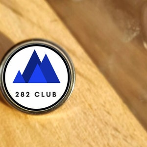 Puede incluir: Un pin metálico redondo con un diseño de montaña azul sobre fondo blanco y el texto "282 CLUB" debajo. El pin está sobre una superficie de madera marrón claro.