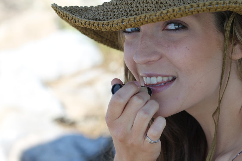 Puede incluir: Una mujer con un sombrero de paja tejido marr&oacute;n se aplica b&aacute;lsamo labial con la mano derecha.