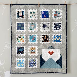 Könnte beinhalten: Eine graue Steppdecke mit dunkelblauem Rand, die quadratische, briefmarkenähnliche Quadrate mit verschiedenen Designs aufweist. Die Steppdecke enthält Bilder von Katzen, Büchern und einem Umschlag mit einem Herzen.