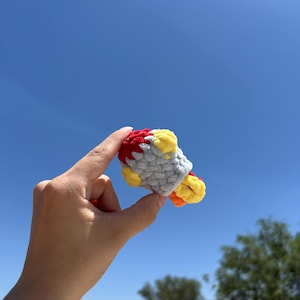 May include: A small, handmade crochet rocket ship in red, gray, yellow, and orange. The toy is held against a bright blue sky, with green trees in the background. The rocket has a textured, plush appearance.
