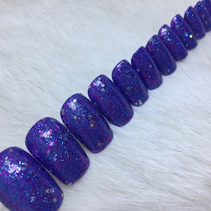 Puede incluir: Un conjunto de diez uñas postizas de color azul y morado con purpurina. Las uñas tienen forma ovalada y un acabado brillante.