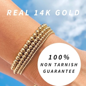 Op de afbeelding: Een close-up van een pols met vier gouden kralen armbanden. De armbanden zijn op elkaar gestapeld en gemaakt van kleine, ronde kralen. De tekst "REAL 14K GOLD" staat bovenaan de afbeelding en "100% NON TARNISH GUARANTEE" staat onderaan de afbeelding.