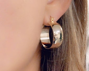 May include: Gold hoop earrings with a thick, rounded band. The earrings are worn in a person's ear.