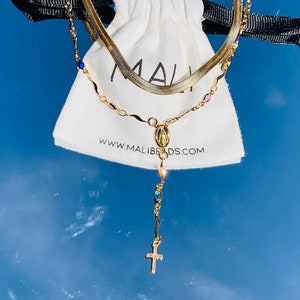 May include: A gold chain necklace with a small cross pendant and a charm of the Virgin Mary. The necklace is layered with a second gold chain. The necklace is displayed on a white pouch with the text "www.malibeads.com".