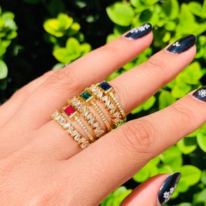 以下が含まれることがあります： 透明な石と色のついた石がついた金の指輪が5つ。指輪は指に重ねられています。石は赤、緑、青、透明です。