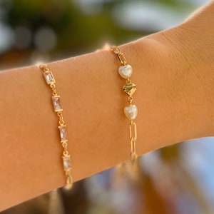 May include: Two gold bracelets on a wrist. One bracelet has a delicate chain with small rectangular clear stones. The other bracelet has a thicker chain with three heart-shaped charms, two gold and one white.