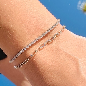 Op de afbeelding: Twee zilveren armbanden worden getoond op een pols. De bovenste armband is een tennisarmband met kleine, heldere stenen. De onderste armband is een schakelarmband. De achtergrond is een heldere blauwe lucht.