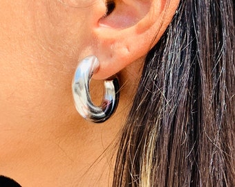 May include: Silver hoop earrings with a thick, rounded design. The earrings are shown on a person's ear.