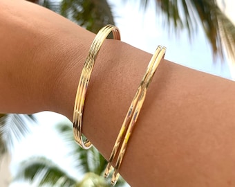 May include: Three gold bangles stacked on a person's wrist. The bangles are thin and have a smooth, polished finish.