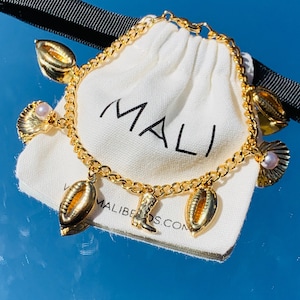 May include: Gold chain bracelet with multiple charms, including seashells, a cowboy boot, and a pearl. The bracelet is in a white drawstring bag with the word "MALI" printed on it.