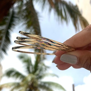 Può includere: Quattro braccialetti rigidi dorati impilati uno sull'altro. I braccialetti sono sottili e hanno una finitura lucida.
