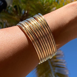 Puede incluir: Una pila de brazaletes de oro y plata, usados en la muñeca de una persona. Los brazaletes están apilados uno encima del otro, creando un look en capas.