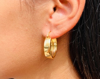 May include: Gold hoop earrings with a thick, smooth, rounded design. The earrings are worn in a person's ear.