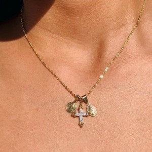 Puede incluir: Un collar de cadena dorada con un pequeño colgante de cruz y dos medallones redondos. La cruz tiene pequeñas piedras transparentes.