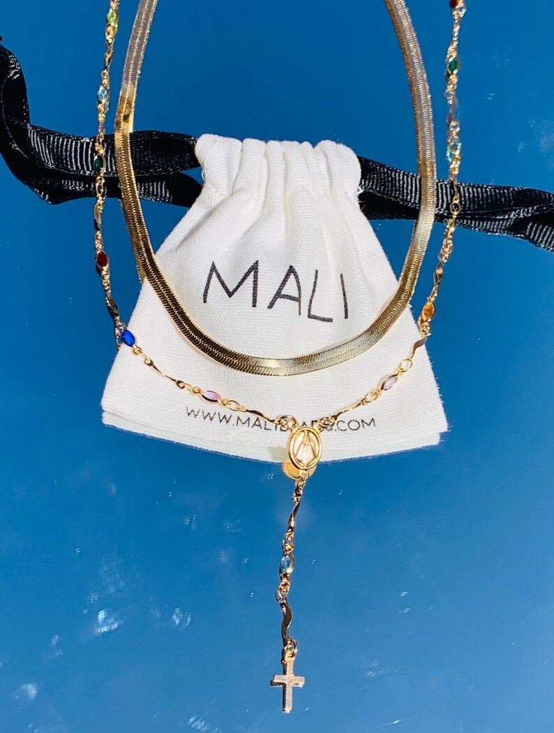 May include: Two gold necklaces, one with a snake chain and the other with colorful beads and a pendant featuring the Virgin Mary and a cross. The necklaces are displayed on a white pouch with the word "MALI" printed on it.
