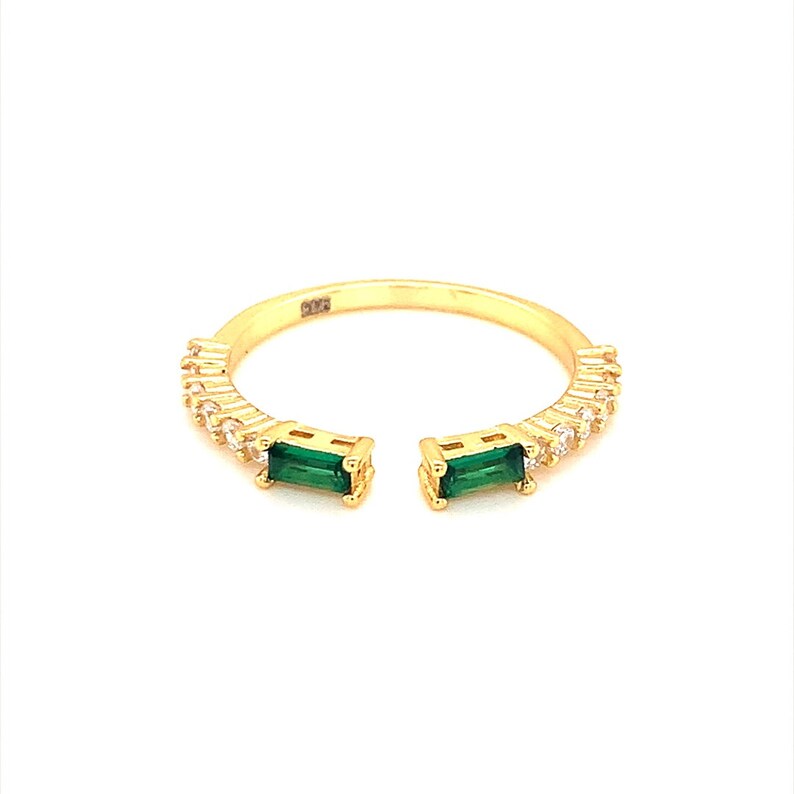 May include: Gold ring with a split band design, featuring two emerald green stones and a row of clear cubic zirconia stones.