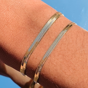 May include: Two gold and silver snake chain bracelets worn on a person's wrist. The bracelets are layered on top of each other.