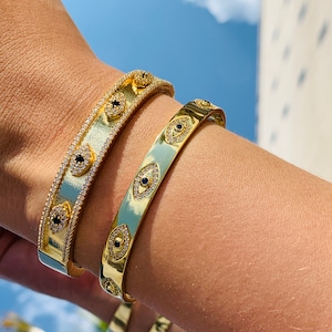 May include: Two gold bracelets with a design of evil eyes with black stones and white diamonds. The bracelets are on a person's wrist.