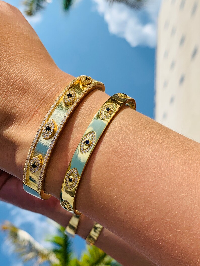 May include: Two gold bracelets with a design of evil eyes with black stones and white diamonds. The bracelets are on a person's wrist.