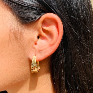 May include: A pair of gold hoop earrings with a slightly curved, rectangular shape. The earrings are worn in the left ear of a person with long, dark hair.