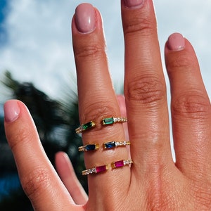 May include: Three gold rings with a single, rectangular, colored gemstone on each. The rings are set with small, clear gemstones around the band. The gemstones are green, blue, and red.