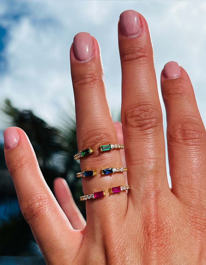 May include: Three gold rings with a single, rectangular, colored gemstone on each. The rings are set with small, clear gemstones around the band. The gemstones are green, blue, and red.