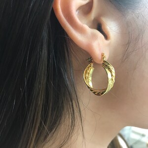 May include: A gold-toned hoop earring featuring a twisted, rope-like texture on one side and a smooth, polished surface on the other. The earring is circular in shape with a hinged clasp mechanism visible at the top, securing it to the earlobe.