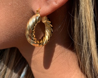 May include: Gold-colored hoop earrings with a twisted rope design. The earrings are a medium size and have a shiny finish. The image shows the earring on a person's ear, highlighting the texture and shape of the jewelry.
