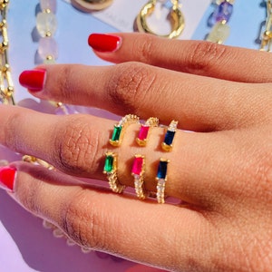 May include: Gold ring with a row of small clear stones and a single emerald green, ruby red, or sapphire blue stone. The ring is shown on a finger.