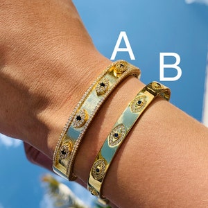 May include: Two gold bracelets with a design of evil eyes set with clear and black stones. The bracelets are on a person's wrist. The bracelet labeled 'A' is wider than the bracelet labeled 'B'.