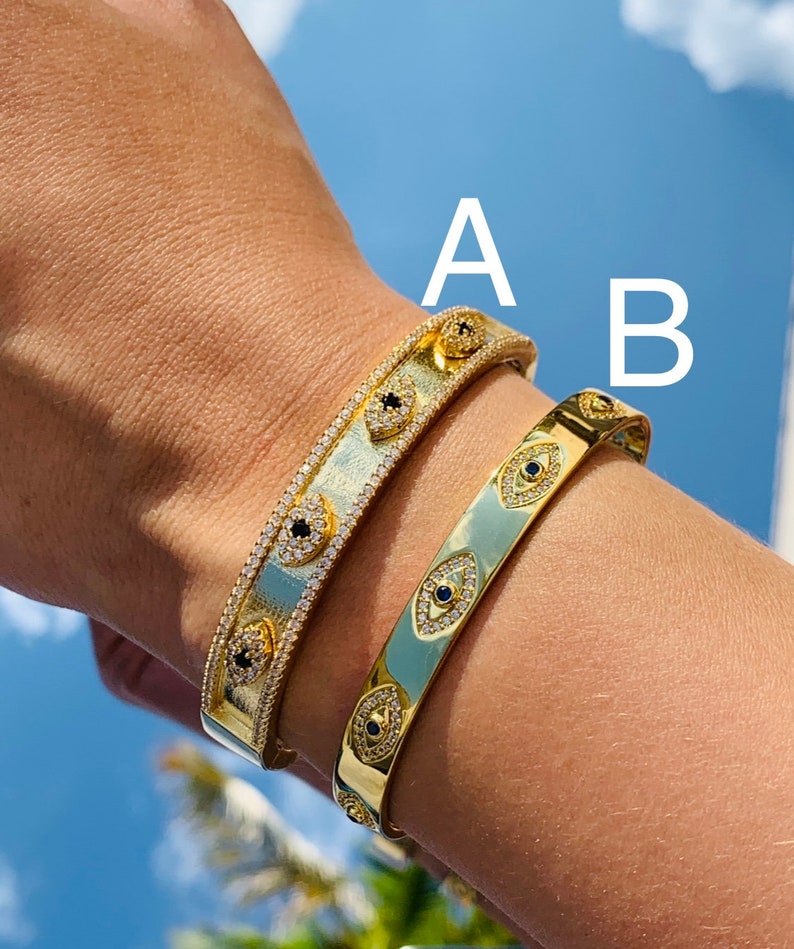 May include: Two gold bracelets with a design of evil eyes set with clear and black stones. The bracelets are on a person's wrist. The bracelet labeled 'A' is wider than the bracelet labeled 'B'.