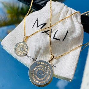 May include: Two gold chain necklaces with circular pendants featuring a prayer inscribed in gold lettering. The necklaces are displayed on a white fabric pouch with the word "MALI" printed on it.
