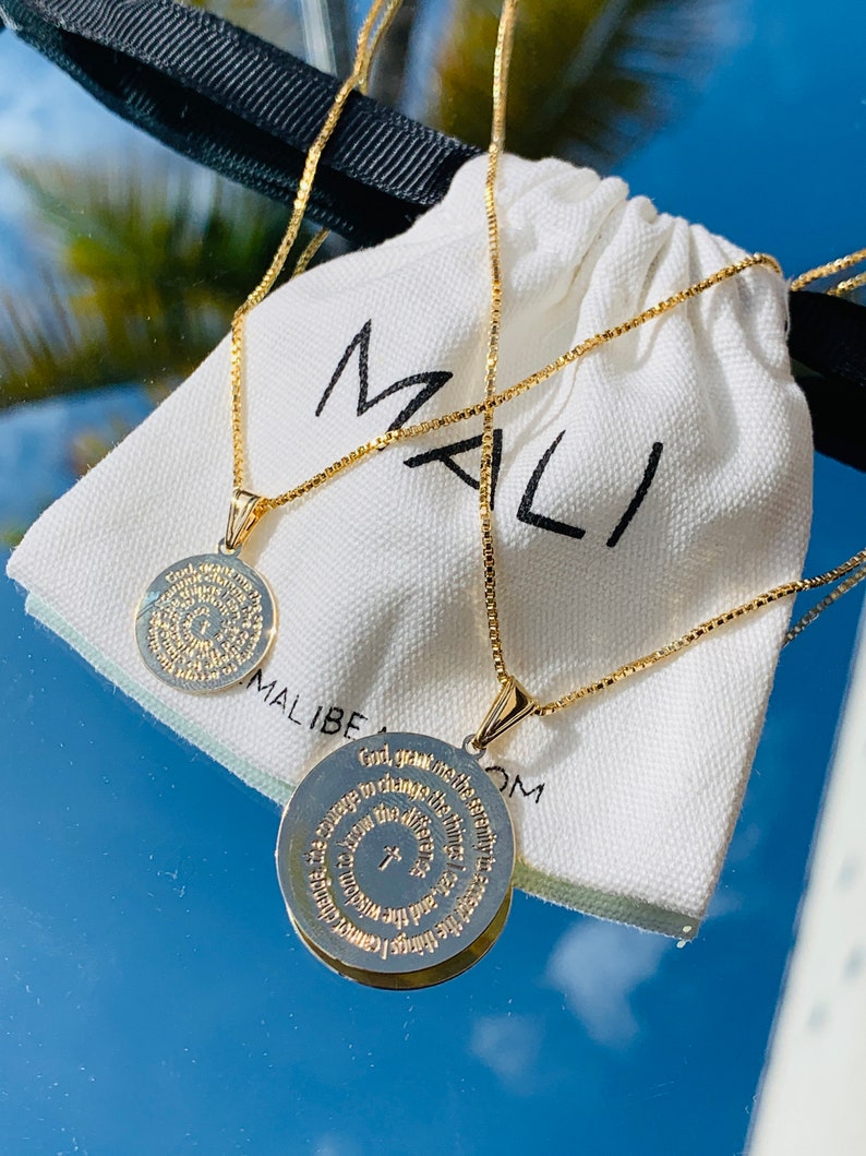 May include: Two gold chain necklaces with circular pendants featuring a prayer inscribed in gold lettering. The necklaces are displayed on a white fabric pouch with the word "MALI" printed on it.