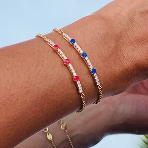 Op de afbeelding: Twee gouden armbanden met een rij heldere stenen en afwisselend rode en blauwe stenen. De armbanden zitten om de pols van een persoon.
