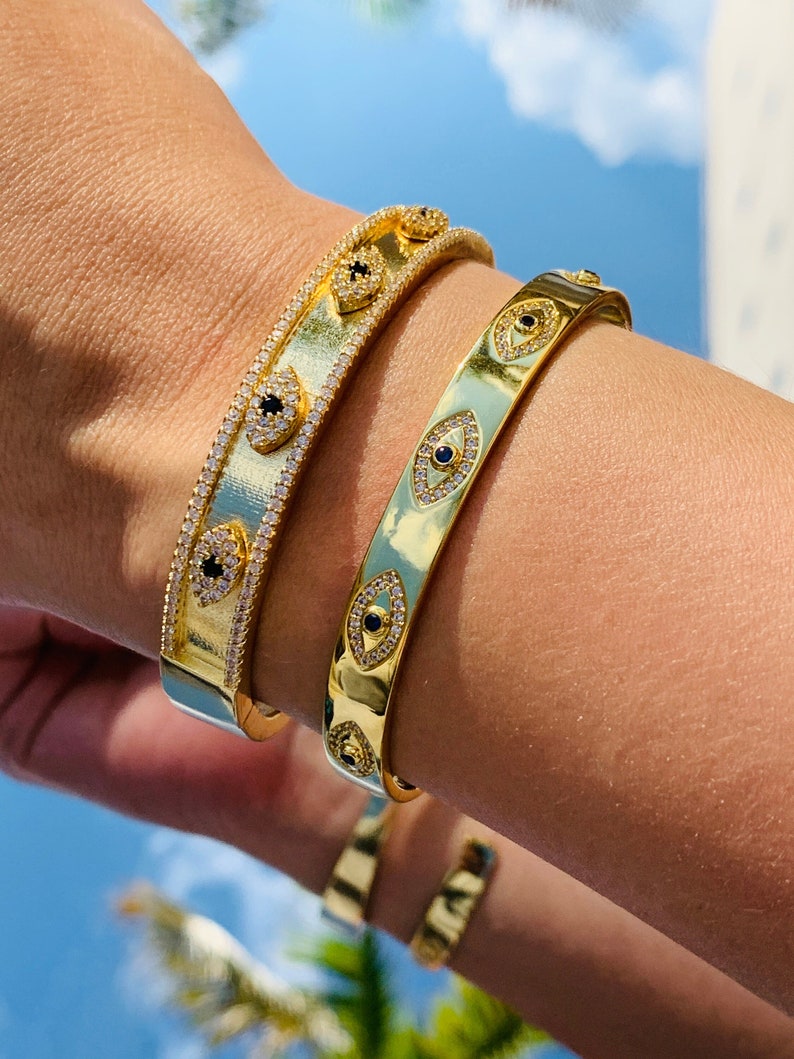 May include: Two gold bracelets with a pave diamond design and black evil eye charms. The bracelets are stacked on a person's wrist.