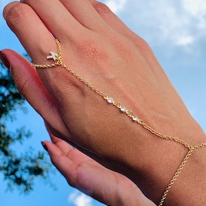 May include: A gold chain bracelet with a small cross charm and clear stones. The bracelet is worn on the wrist and hand, with the chain extending from the ring finger to the wrist.