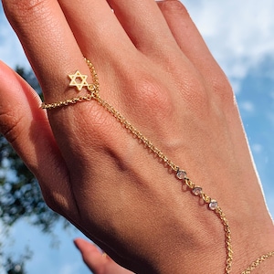 May include: A gold chain bracelet with a star of David charm and small clear stones. The bracelet is worn on a hand with the chain extending up the arm.