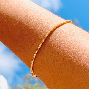 Op de afbeelding: Een gouden slangenketting armband gedragen op de arm van een persoon tegen een blauwe lucht achtergrond.